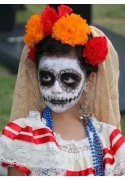 A young girl wearing Día de Muertos skull face paint, a flower crown, and traditional clothing with bright colors and beads.