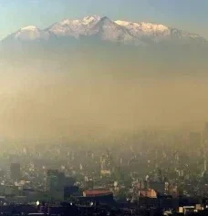Panoramic view of Mexico City beneath a dense layer of air pollution, with mountains rising above the haze in the background, illustrating how the surrounding geography can contribute to the accumulation of smog over the city.